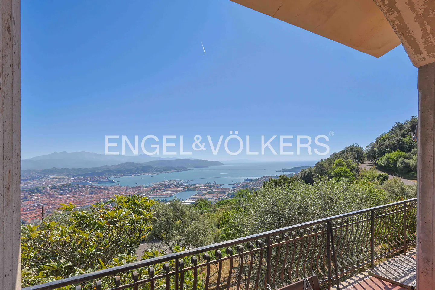 View from a balcony overlooking a city, harbor, and mountains under a blue sky. The balcony has a black metal railing and a red tile floor. Trees and greenery are visible in the foreground.