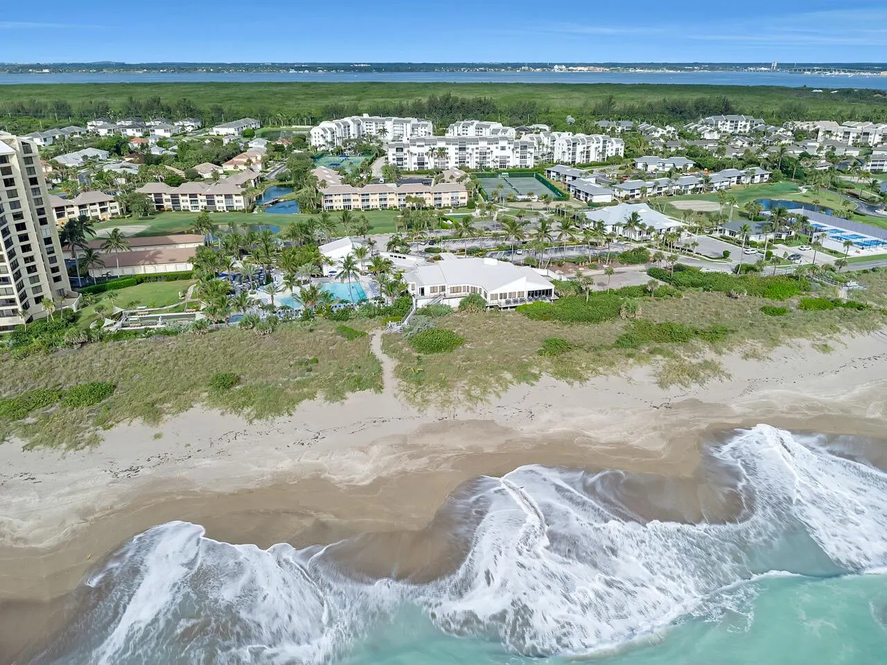 Aerial view of a beach with white waves, sand, and a resort with white buildings, pools, and palm trees.