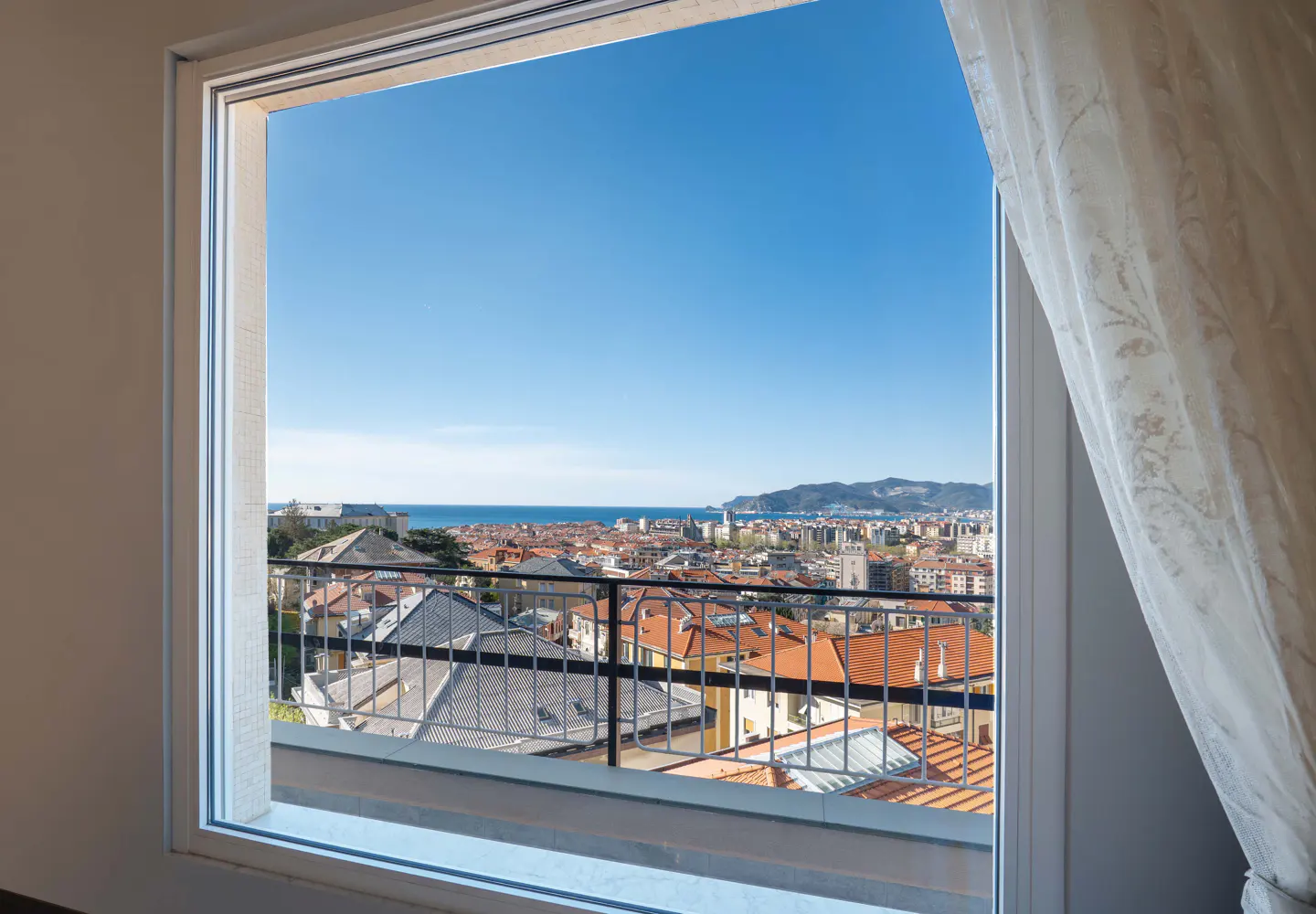 View from a window with white frame and curtain, overlooking a city with red rooftops, the sea, and mountains under a clear blue sky.