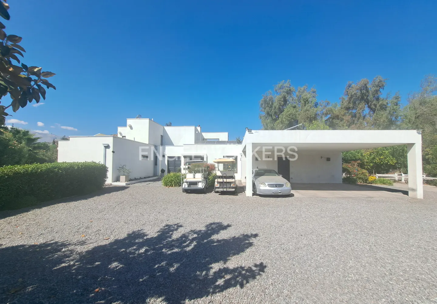 Modern white house with a gravel driveway, two golf carts, and a silver car under a carport on a sunny day.