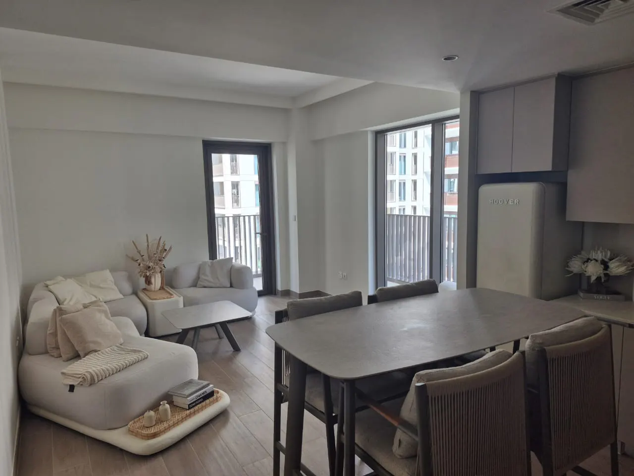 A modern, open-concept living space with a gray dining table, white sofa, and balcony doors. A vintage-style refrigerator is visible in the kitchen area.