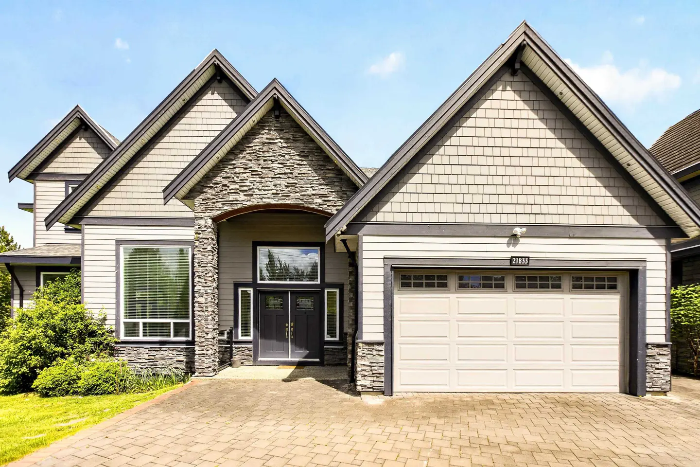 Two-story house with gray siding, stone accents, and a white garage door under a blue sky.