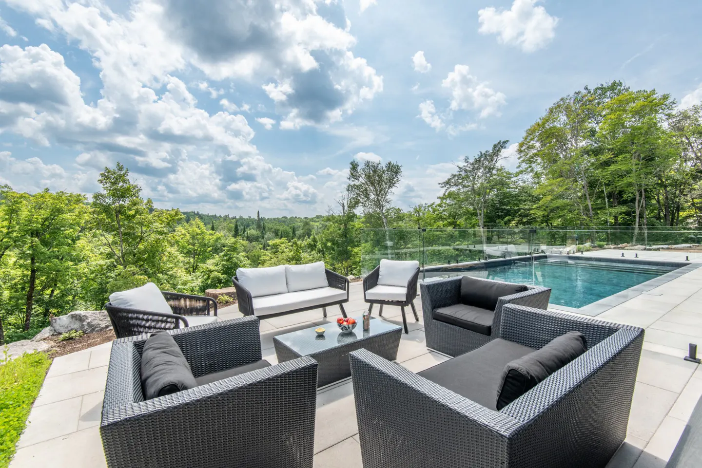 Outdoor patio with black wicker furniture, white cushions, and a glass-top table. A pool is visible behind a glass fence, with trees and a cloudy sky in the background.