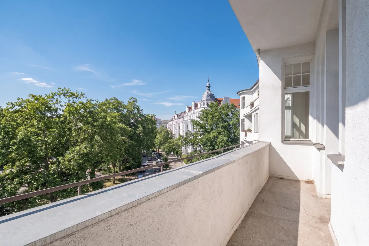 View from a white balcony overlooking a tree-lined street with white buildings and a blue sky.