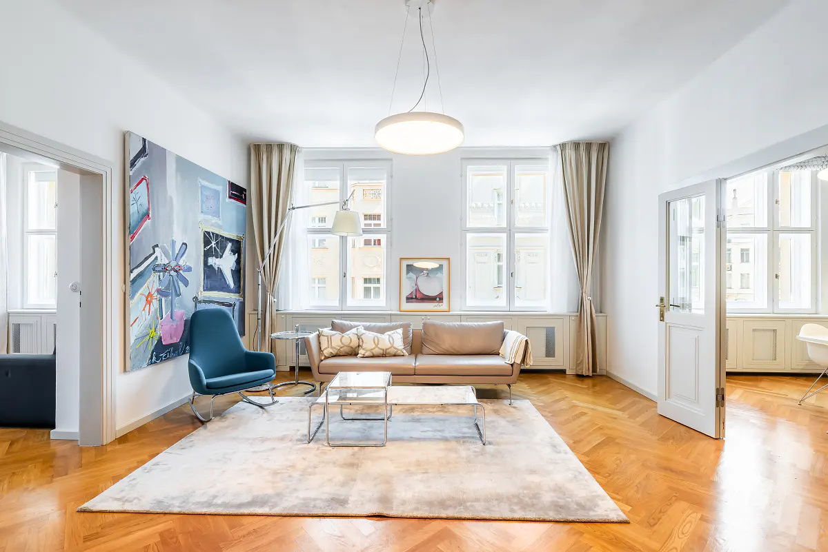 Bright living room with herringbone wood floors, a beige sofa, a blue chair, and a large abstract painting. Natural light streams through the windows.