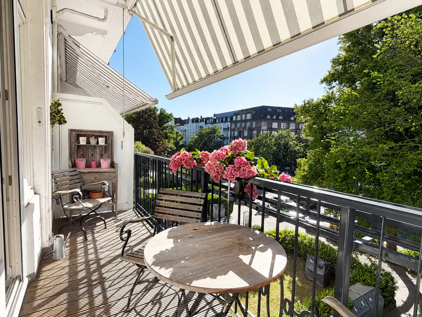 A sunny balcony with a round wooden table, chairs, and pink flowers on the railing overlooking a green cityscape.