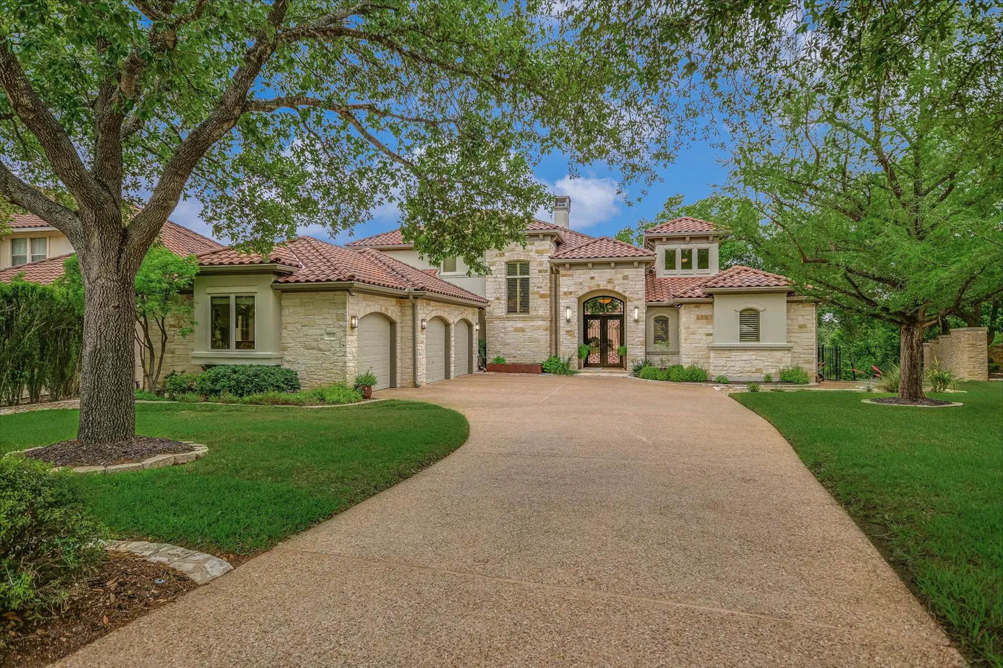 A large, two-story stone house with a red tile roof and a long driveway. Green trees frame the house.