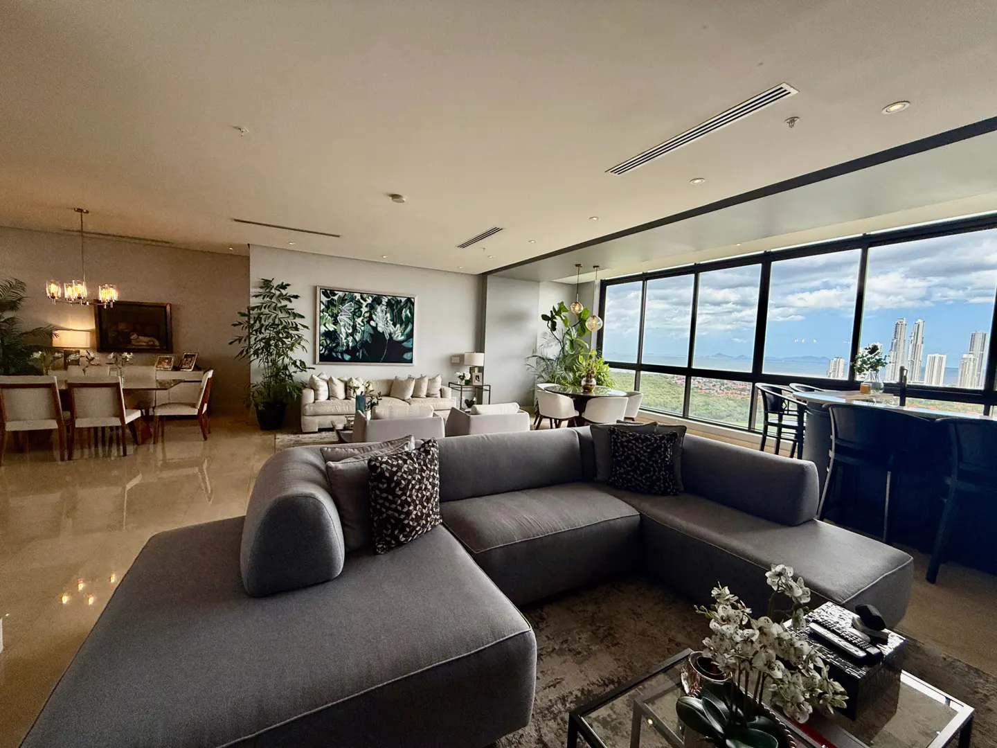 A modern living room with a gray sectional sofa, dining table, and a large window with a city view.