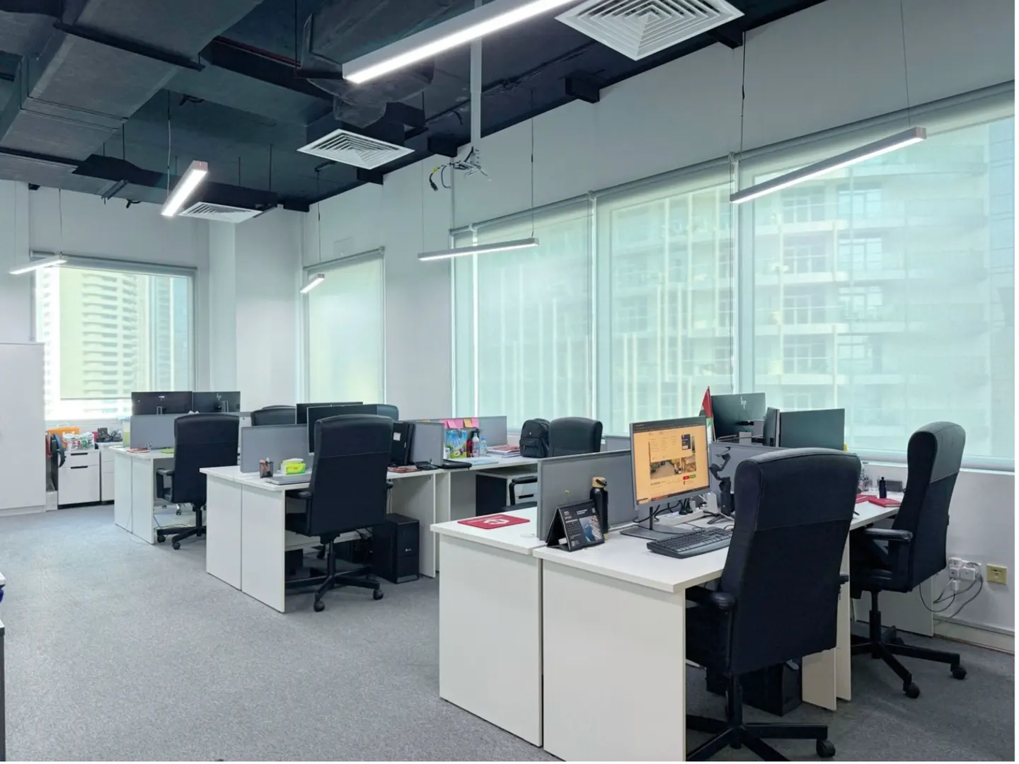 Bright office space with white desks, black chairs, and computers. Large windows offer a city view. Overhead lighting illuminates the workspace.