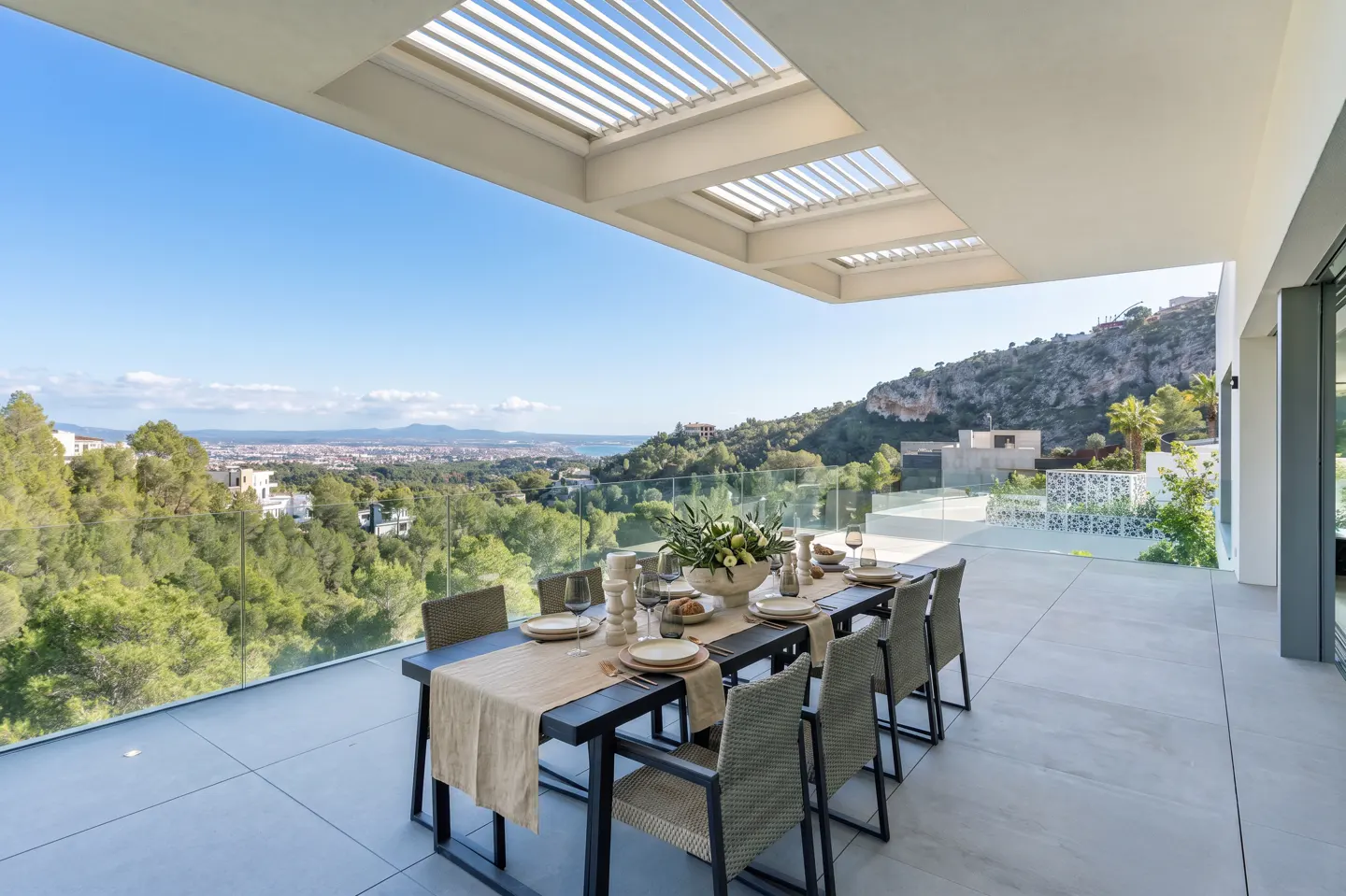 Outdoor dining area with a long table set for a meal, chairs, and a scenic view of trees and a city.