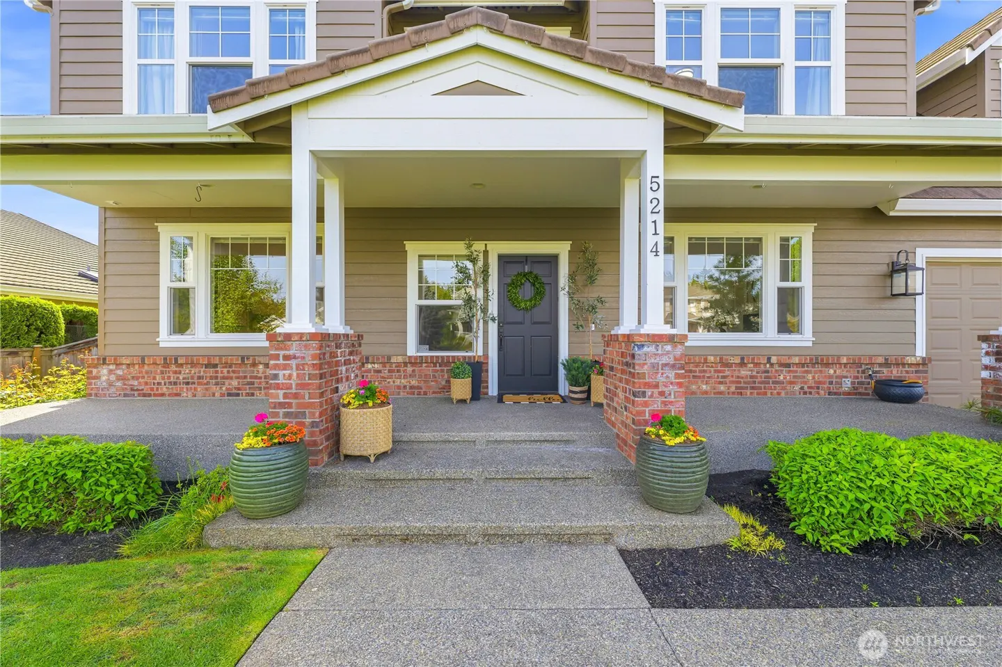 Front view of a two-story house with a gray door, wreath, brick accents, and flower pots on the porch.