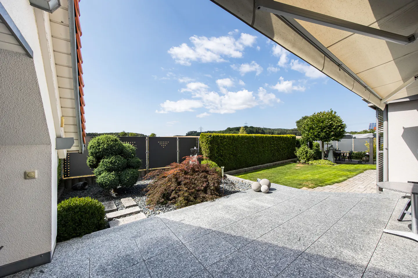 A patio with gray stone tiles leads to a green lawn and garden with a black fence and trimmed hedges under a blue sky.