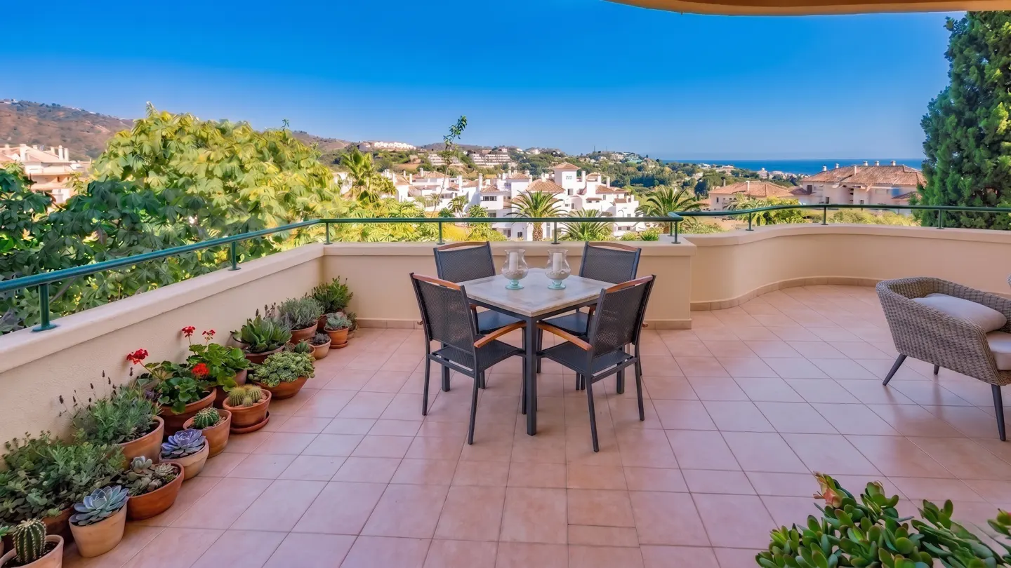 Balcony with table, chairs, and potted plants overlooking a cityscape and ocean under a clear blue sky.