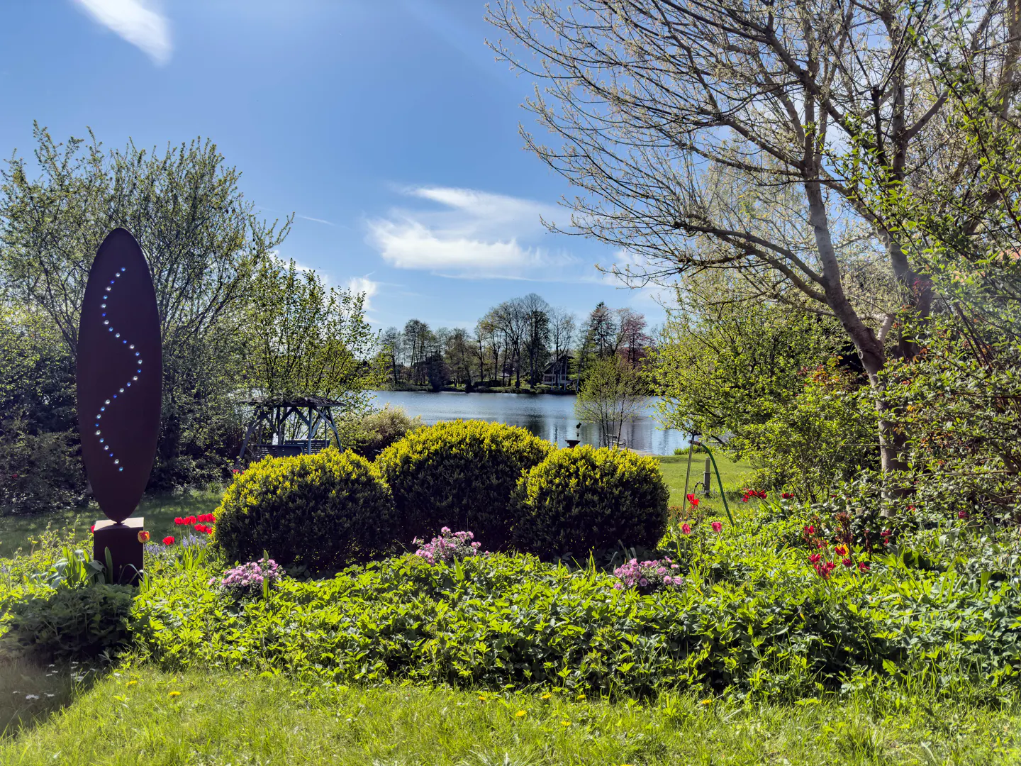 A garden with green bushes, flowers, and a metal sculpture, with a lake and trees in the background under a blue sky.