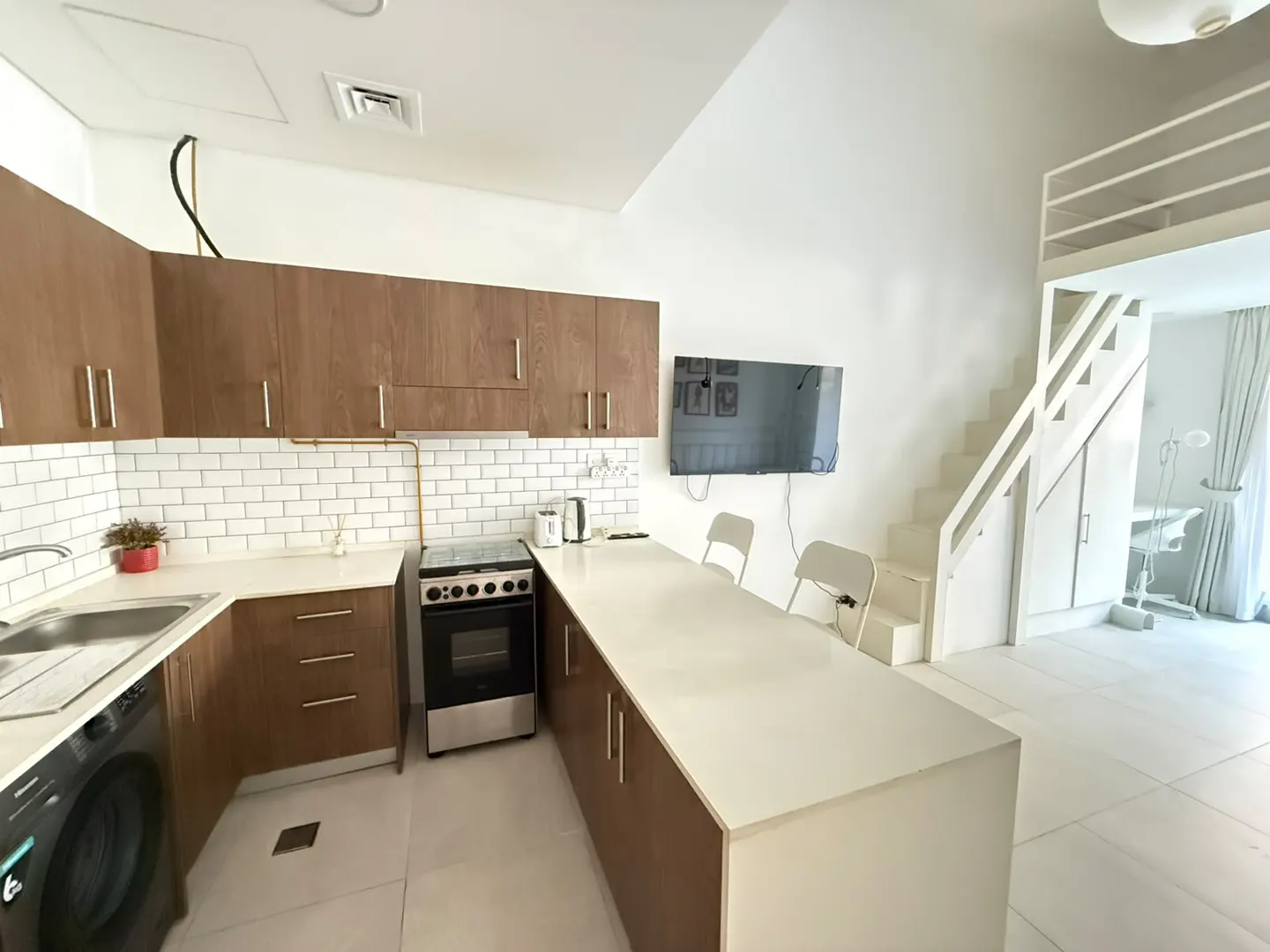 A modern kitchen with brown cabinets, white countertops, and a breakfast bar. A staircase leads to a loft area. A TV is mounted on the wall.