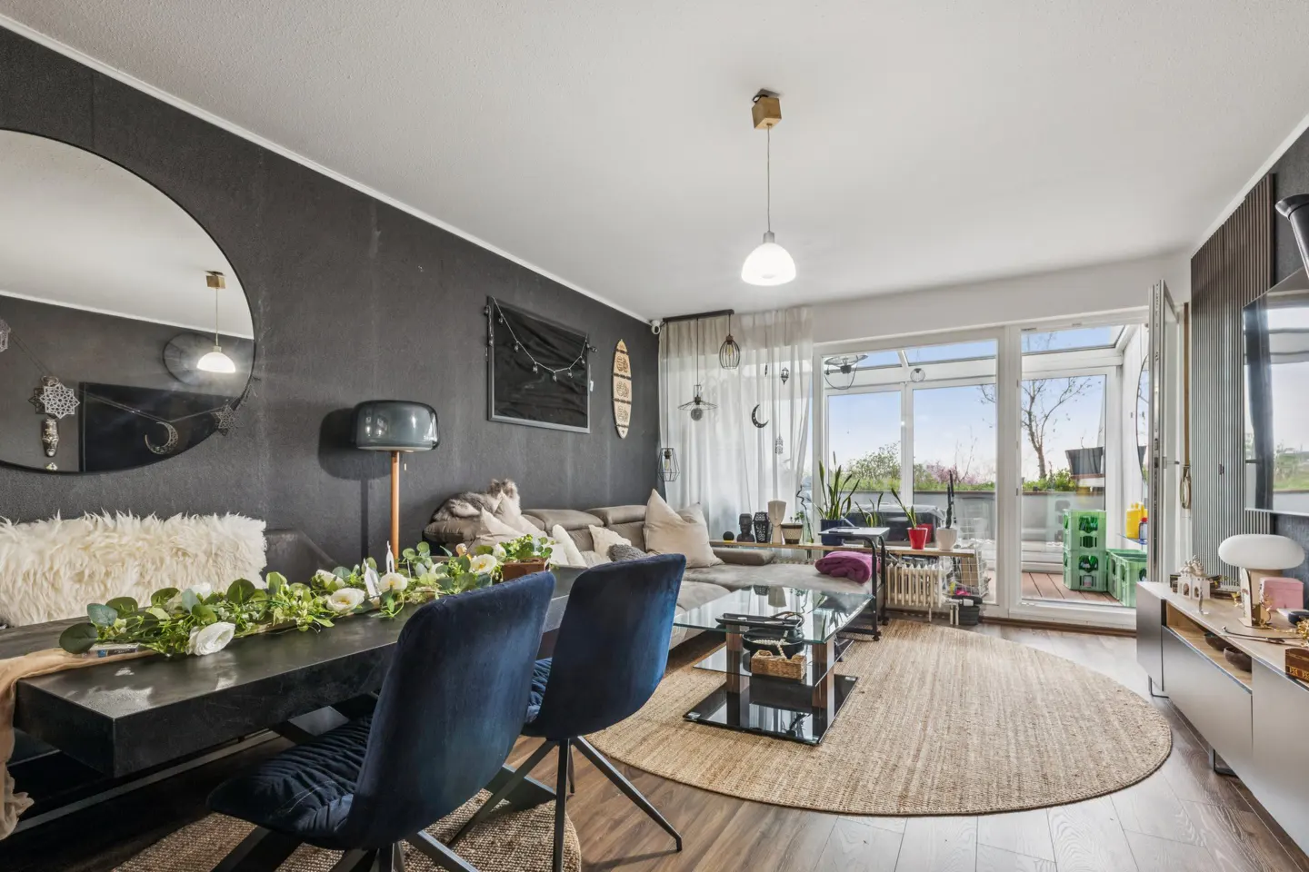 A modern living room with a dark accent wall, dining table with blue chairs, and a balcony with a view.