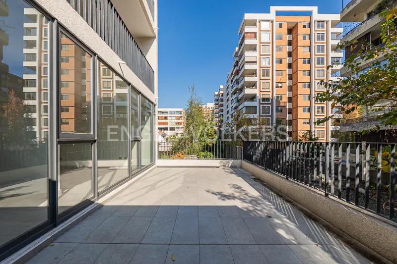 A bright, sunny balcony with gray tile flooring and a black metal railing overlooks apartment buildings and trees.