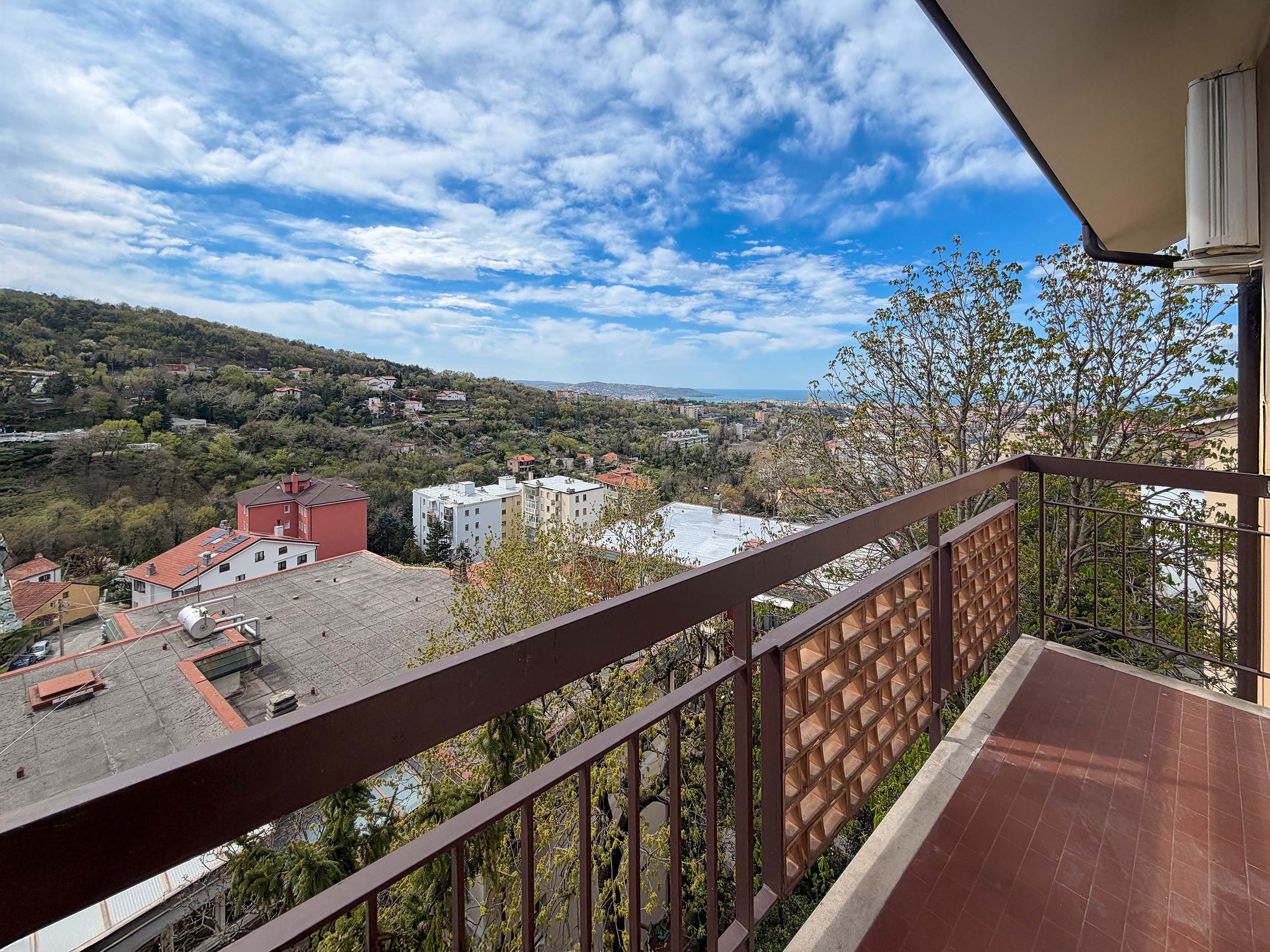 View from a balcony with brown railings overlooking a town with green hills and a blue sky with white clouds.