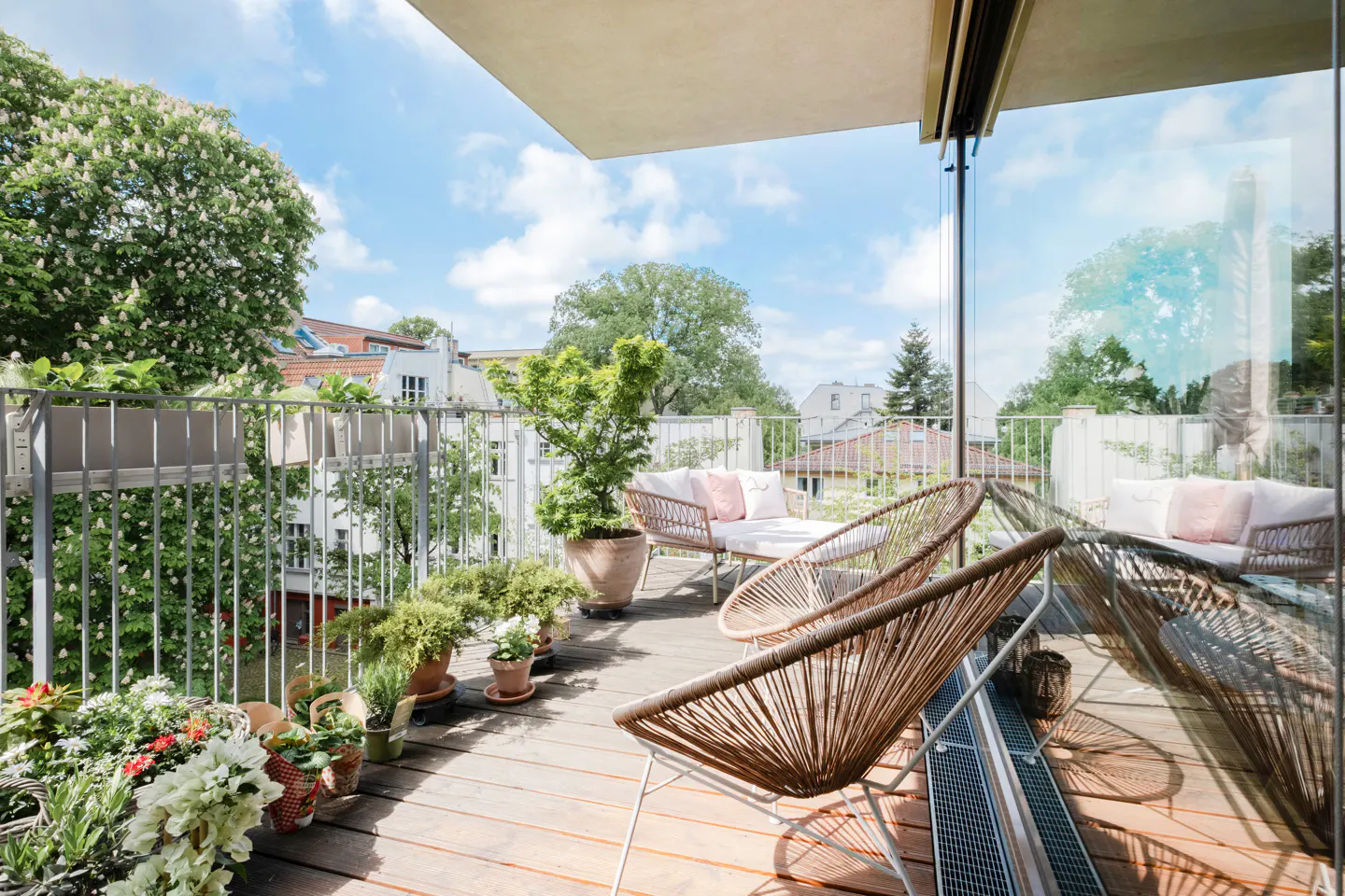 Sunny wooden balcony with wicker chairs, potted plants, and a view of trees and buildings under a blue sky.