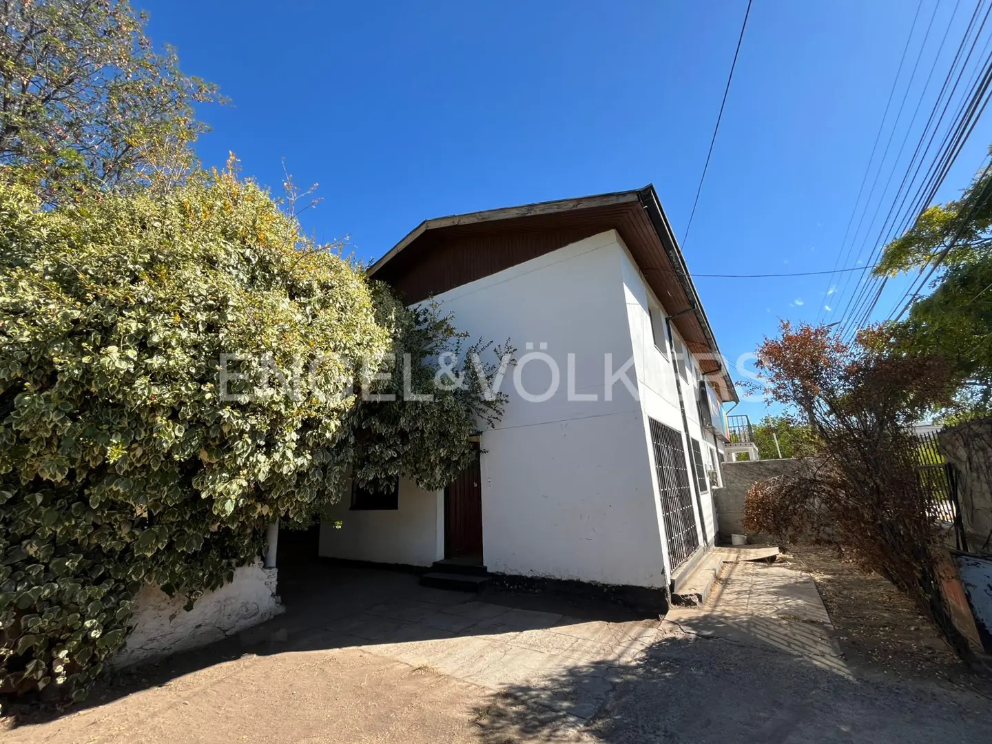 Two-story white house with brown trim, partially obscured by lush green foliage under a clear blue sky. Engel & Völkers logo visible.