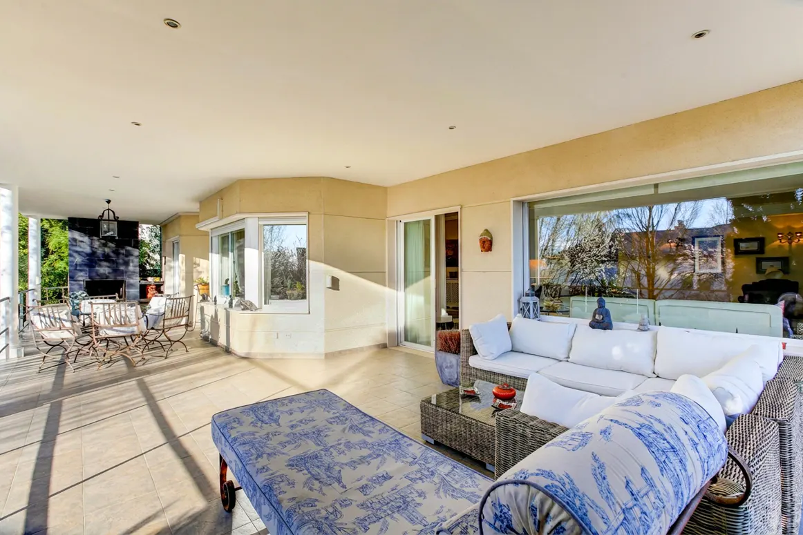 Covered patio with outdoor furniture. A white sofa, wicker table, and blue chaise lounge sit on the tile floor. A dining set is visible in the background.