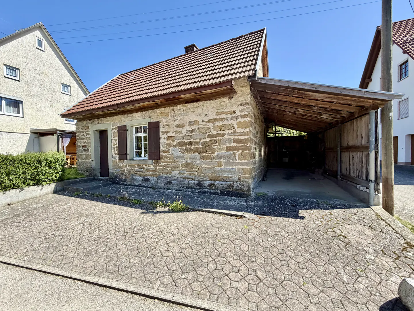 Exterior view of a one-story stone house with a red tile roof and a wooden carport on a sunny day.