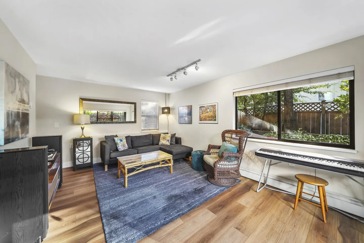 Living room with hardwood floors, a gray sectional sofa, a blue rug, and a wicker chair. A piano sits by the window.