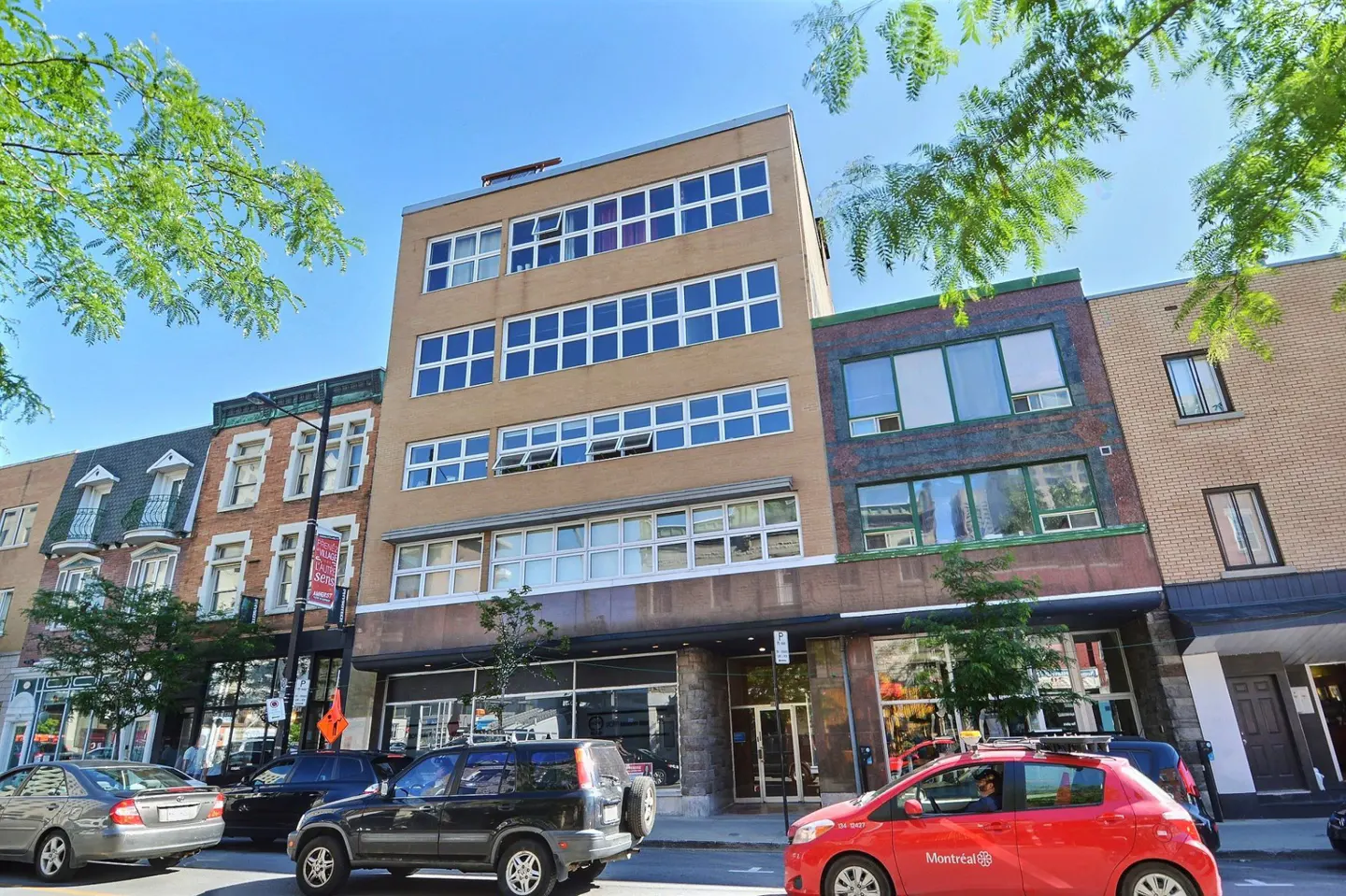 Street view of a tan building with white-framed windows, cars parked in front, and green trees.