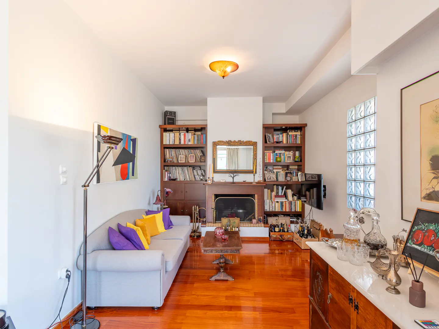 Living room with hardwood floors, a grey sofa with colorful pillows, a fireplace, bookshelves, and a glass block wall.