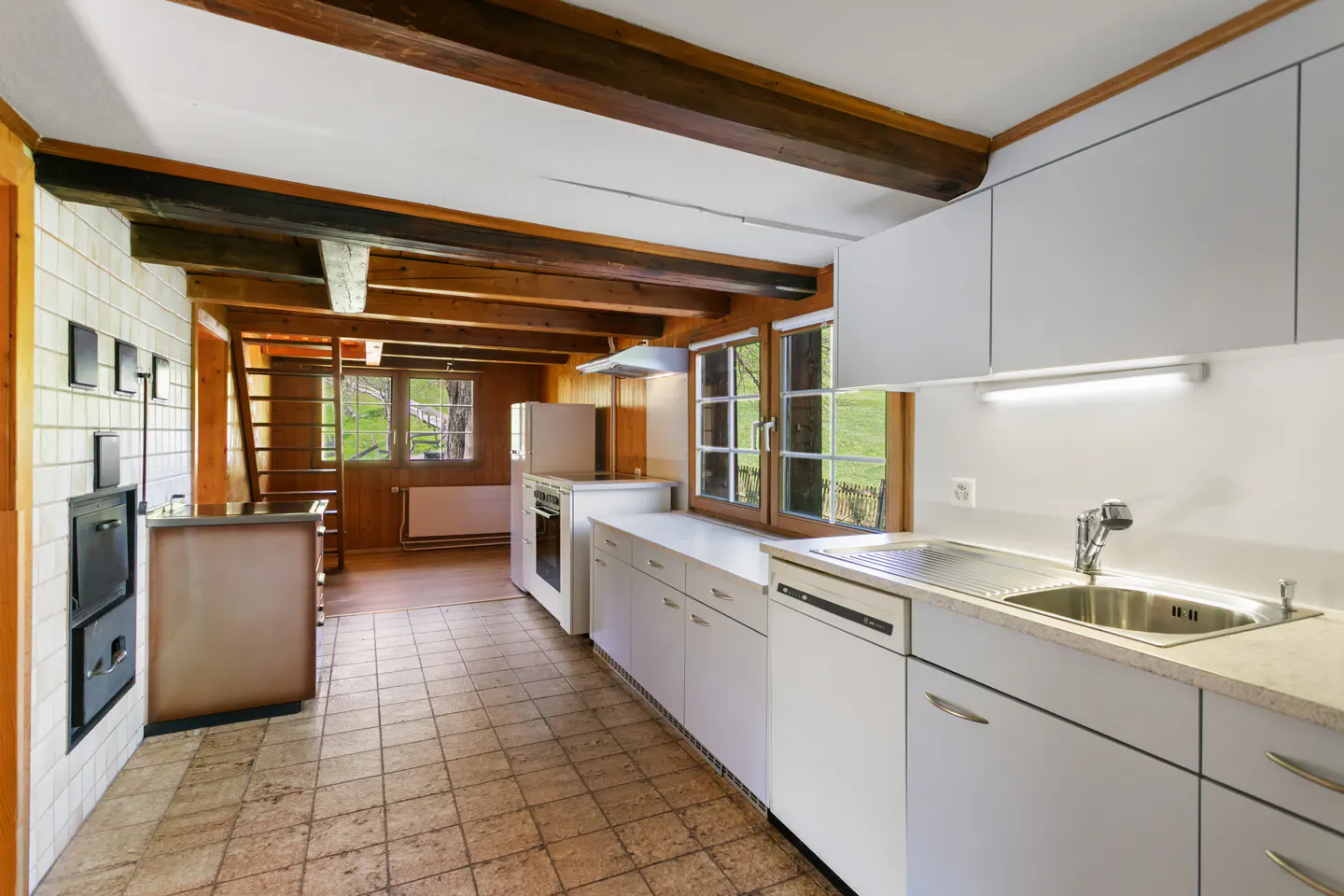 A bright kitchen with white cabinets, a stainless steel sink, and a view of a green field through the window. The kitchen has wooden beams and a tiled floor.