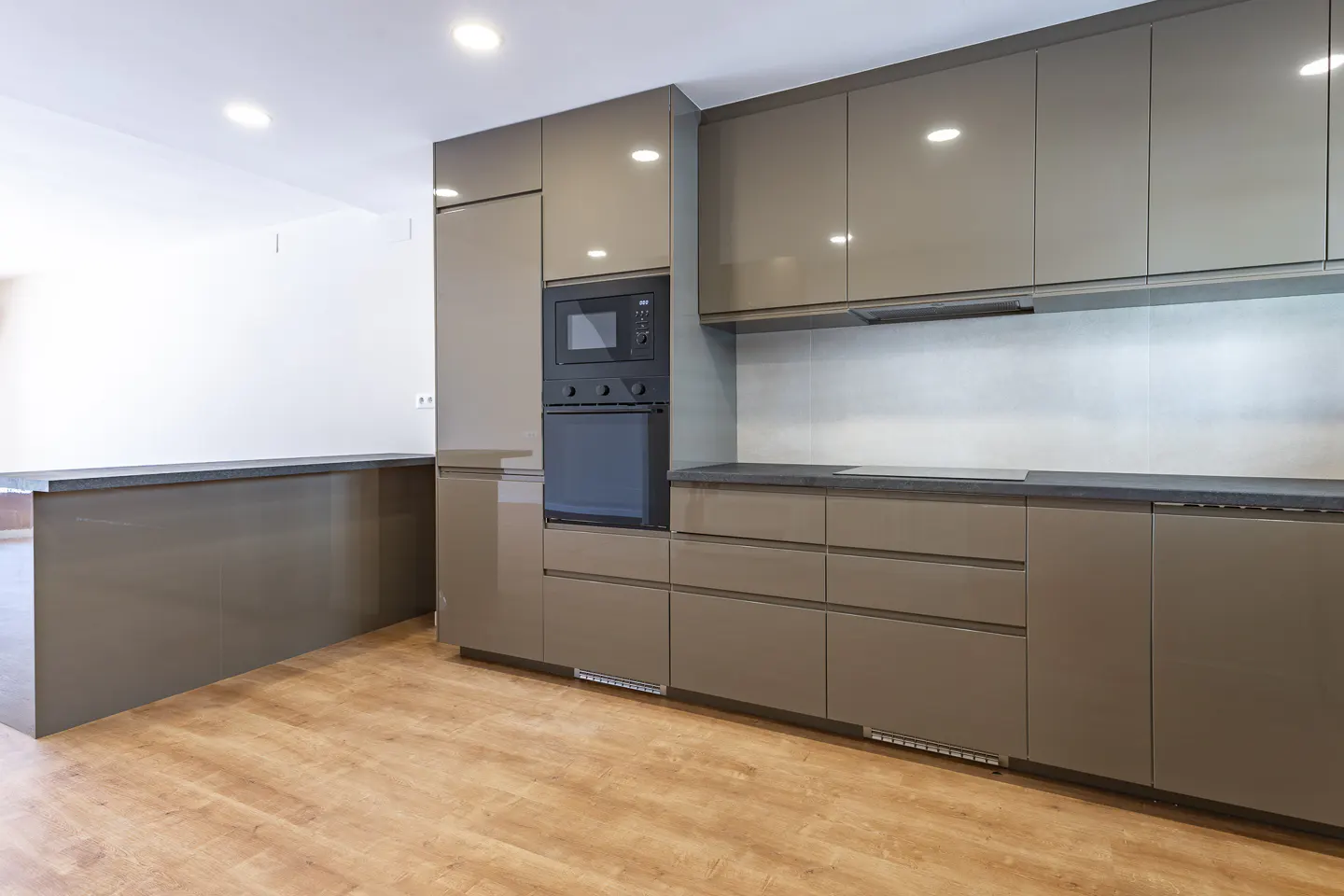Modern kitchen with glossy taupe cabinets, black built-in oven and microwave, dark countertops, and light wood floors.