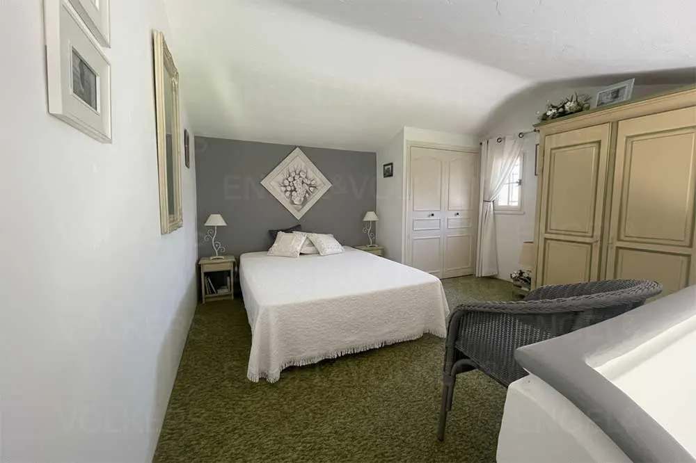 Bedroom with a white bed, gray accent wall, and green carpet. Wardrobe and wicker chair are also visible.