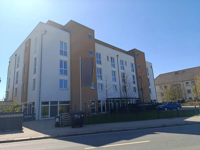 Modern apartment building with white and brown facade, multiple windows, and a blue car parked nearby on a sunny day.