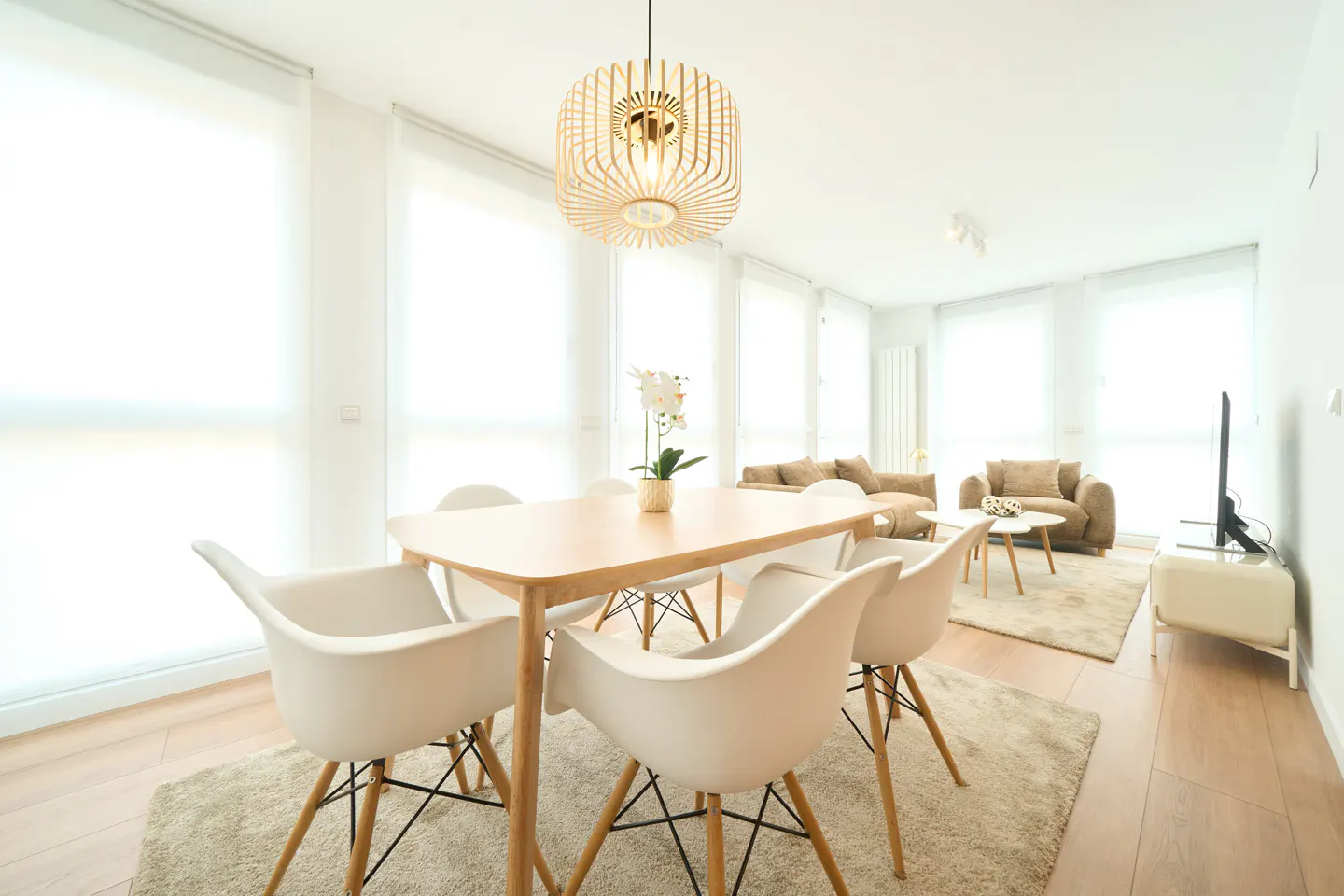 Bright, modern apartment with a light wood dining table, white chairs, and a unique wooden pendant light. Living room with beige sofa in background.
