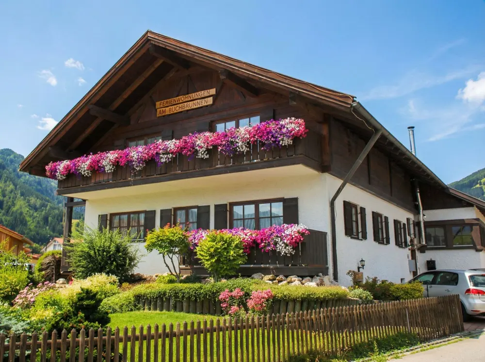 Two-story house with dark wood trim, white walls, and flower boxes on balconies. A wooden fence surrounds a garden with green bushes.