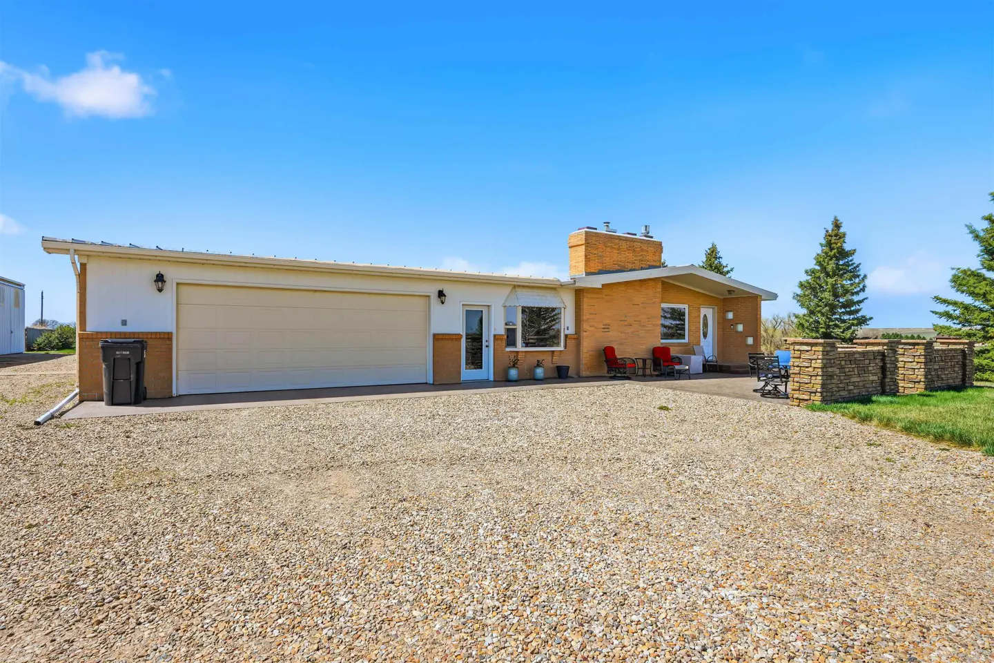 Single-story home with a large garage, brick chimney, and gravel driveway under a clear blue sky.