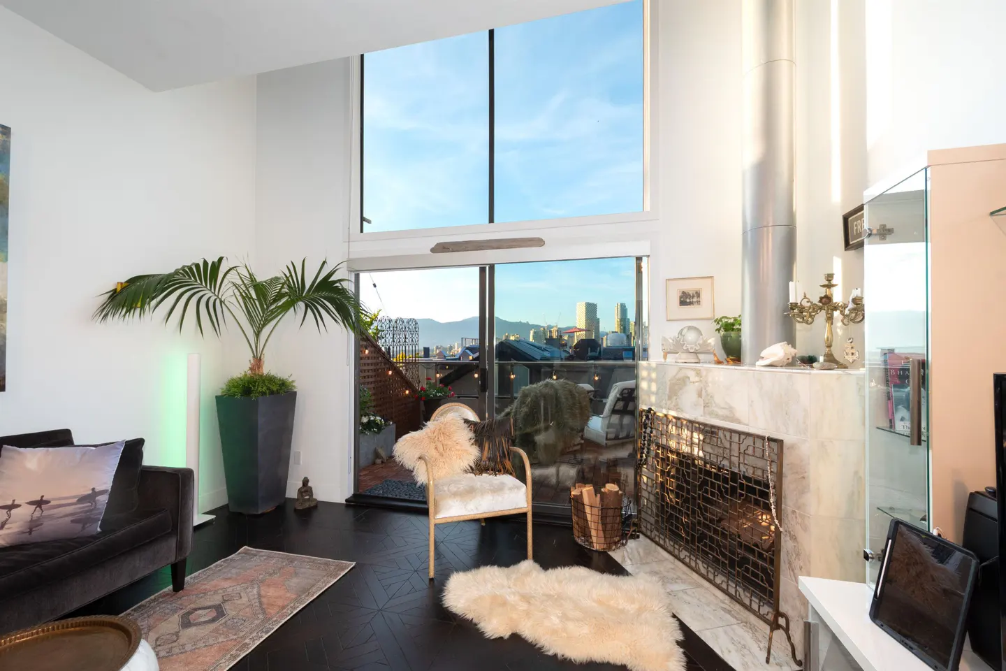 Bright living room with a fireplace, dark wood floors, and a large window overlooking a city skyline. A palm tree and a fur-covered chair add texture.