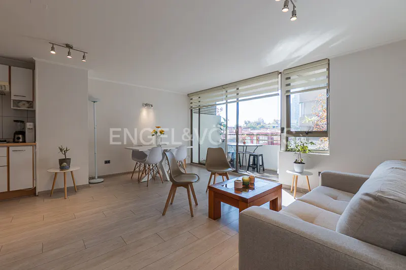 Bright living room with a gray sofa, wood coffee table, and dining area with white table and chairs. Balcony with city views.