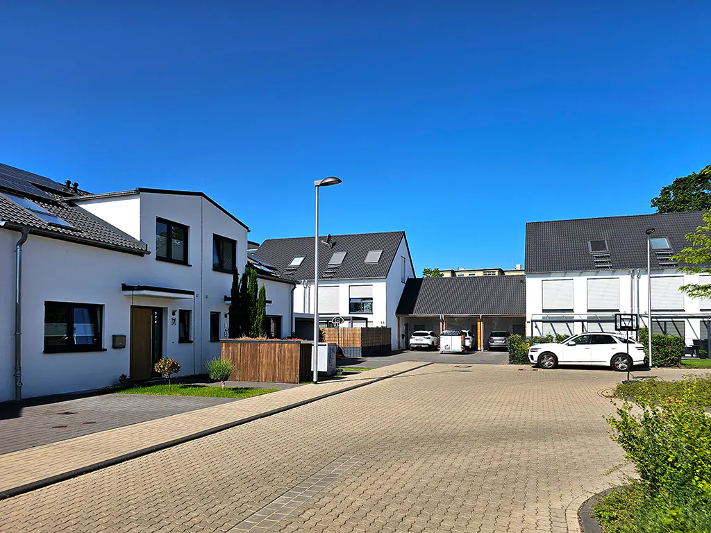 Modern white townhouses with dark roofs and a paved courtyard under a clear blue sky, featuring a parked white car.
