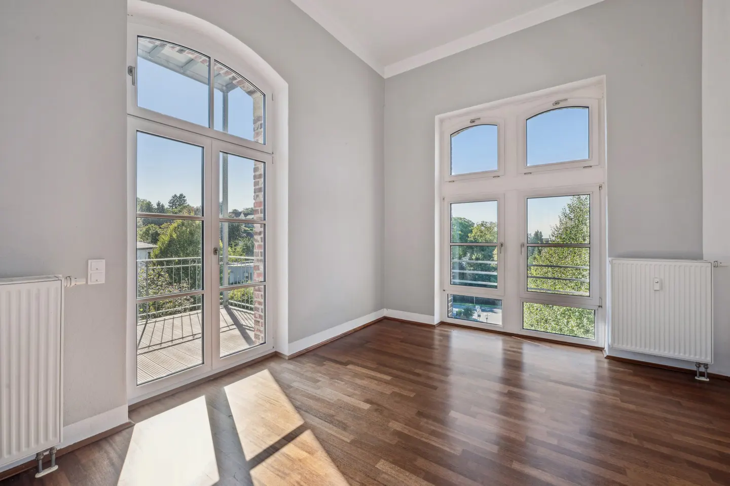 Bright, empty room with hardwood floors, light gray walls, and two large white windows with views of trees and a balcony.