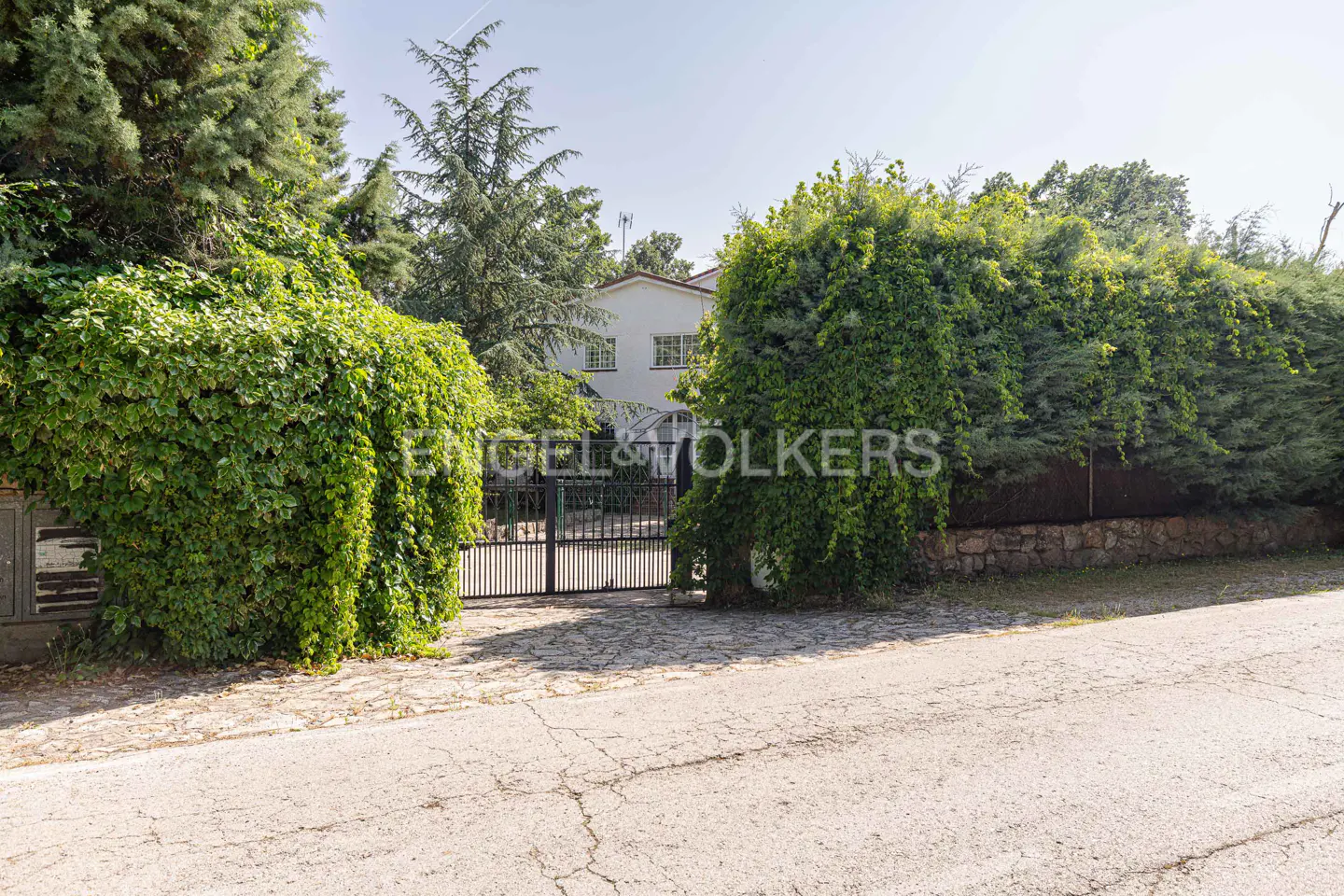 Gated entrance to a white two-story house, surrounded by green trees and bushes. The Engel & Volkers logo is superimposed on the gate.