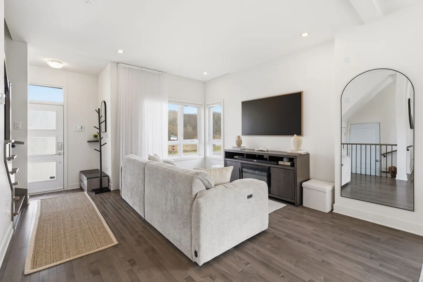 Bright living room with a gray sofa, dark wood floors, and white walls. A large TV is mounted above a dark wood console. A mirror reflects the staircase.