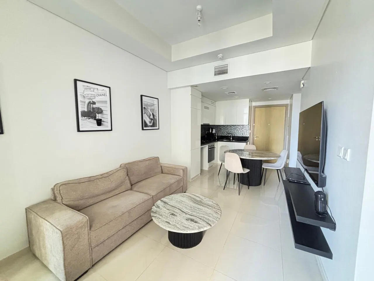 Bright apartment interior with a beige sofa, marble coffee table, and round dining table with white chairs. Black and white art adorns the white walls.