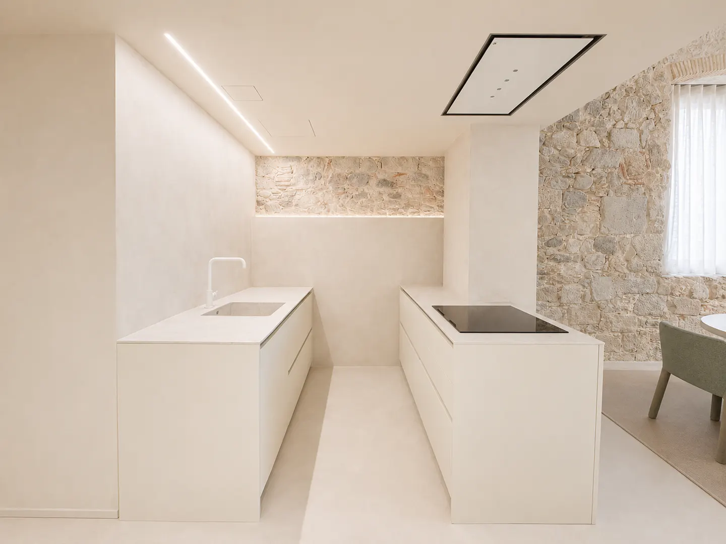 Minimalist kitchen with white cabinets, stone wall, and modern appliances. A sink and stovetop are visible. Natural light filters through a window.