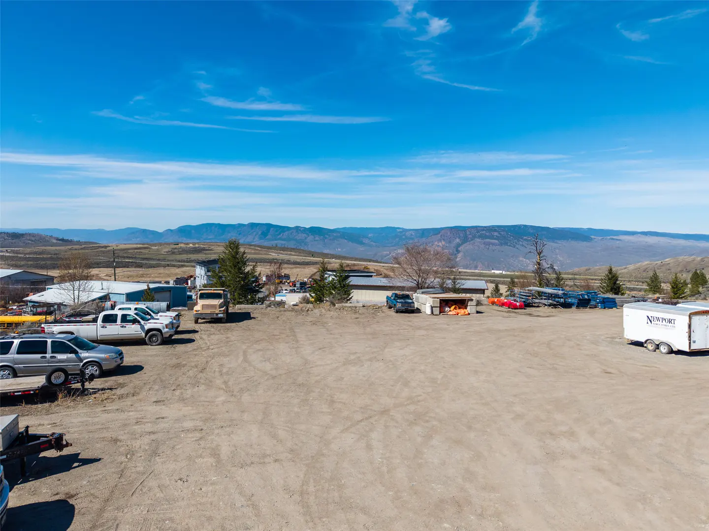Wide shot of a dirt lot with parked vehicles, buildings, and mountains under a blue sky.