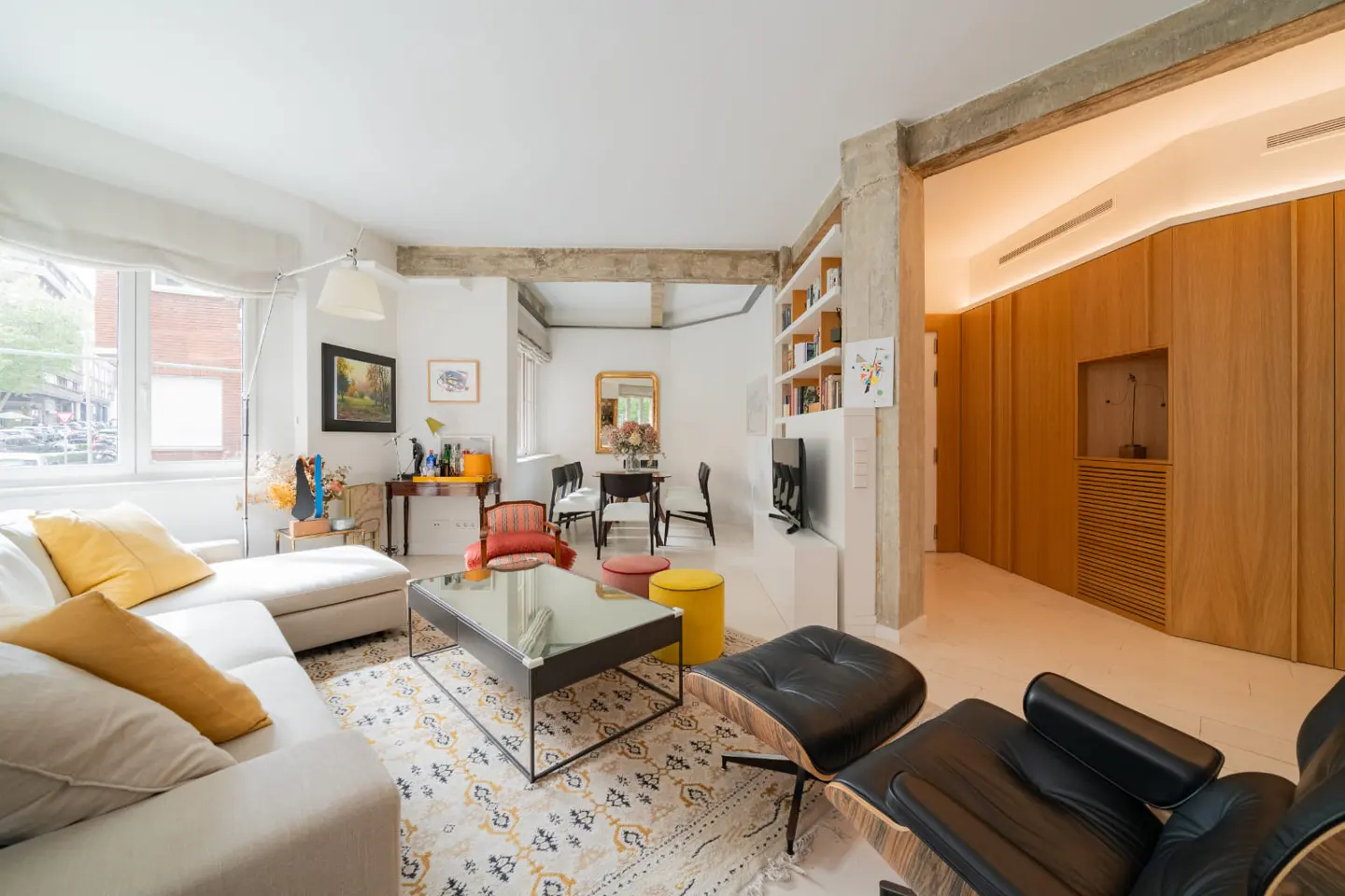 Bright, modern living room with white walls, concrete beams, and light wood accents. A white sofa, black leather chair, and patterned rug furnish the space.