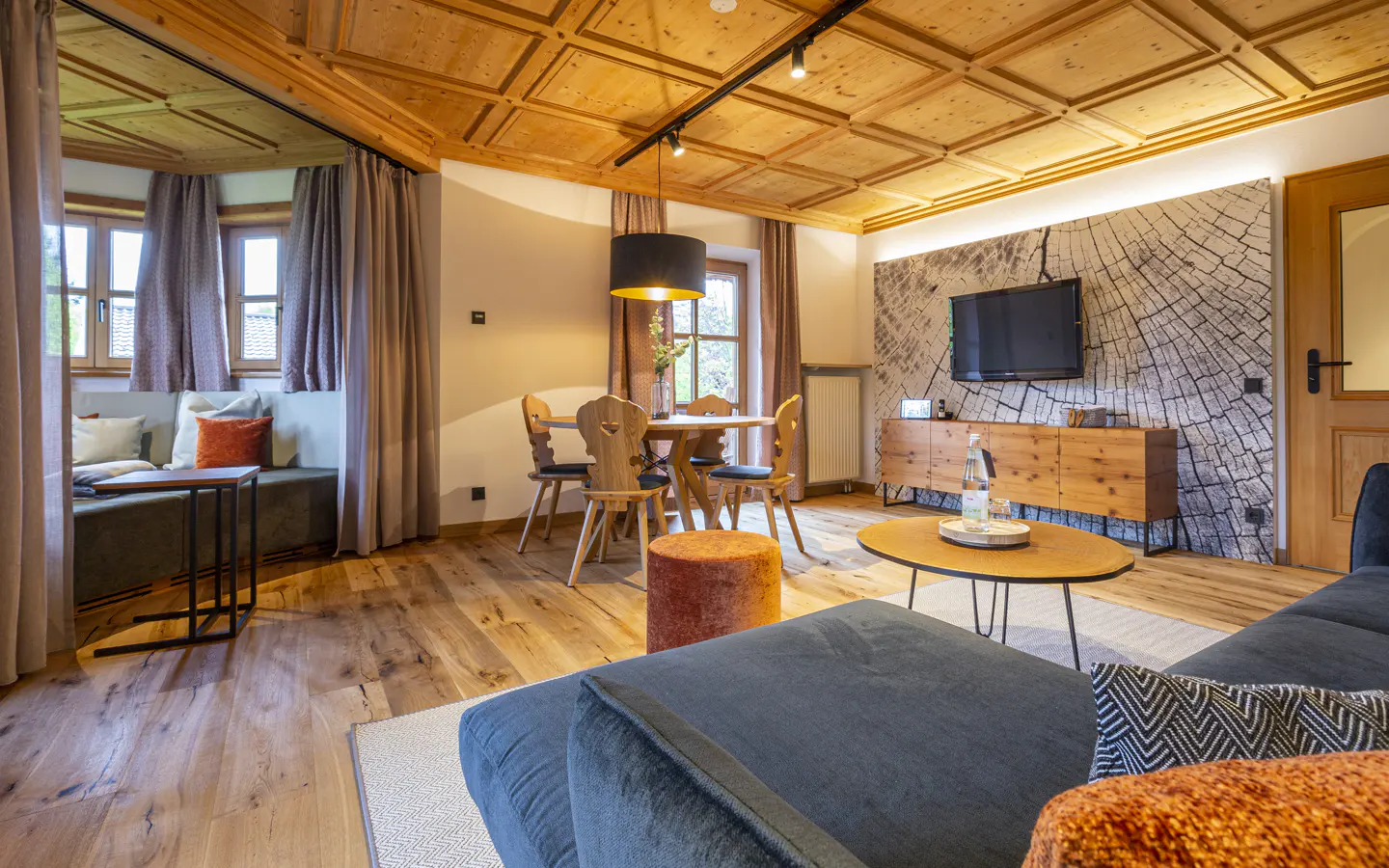 Cozy living room with wood-paneled ceiling, blue sofa, round table, and dining set. A tree-ring wallpaper adds a rustic touch.