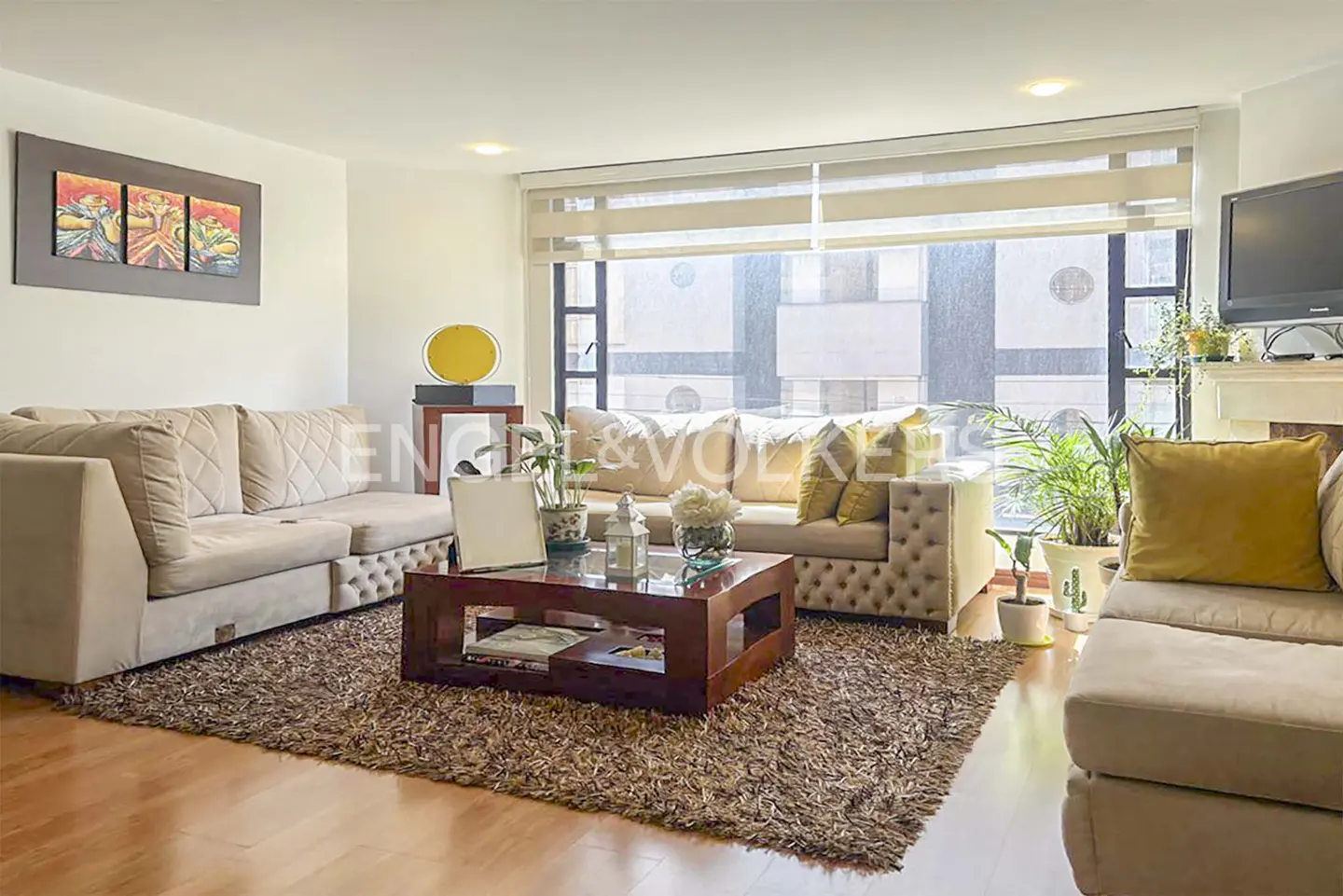 Living room with beige sofas, a dark wood coffee table, and a large window with blinds. A brown rug covers the wood floor.