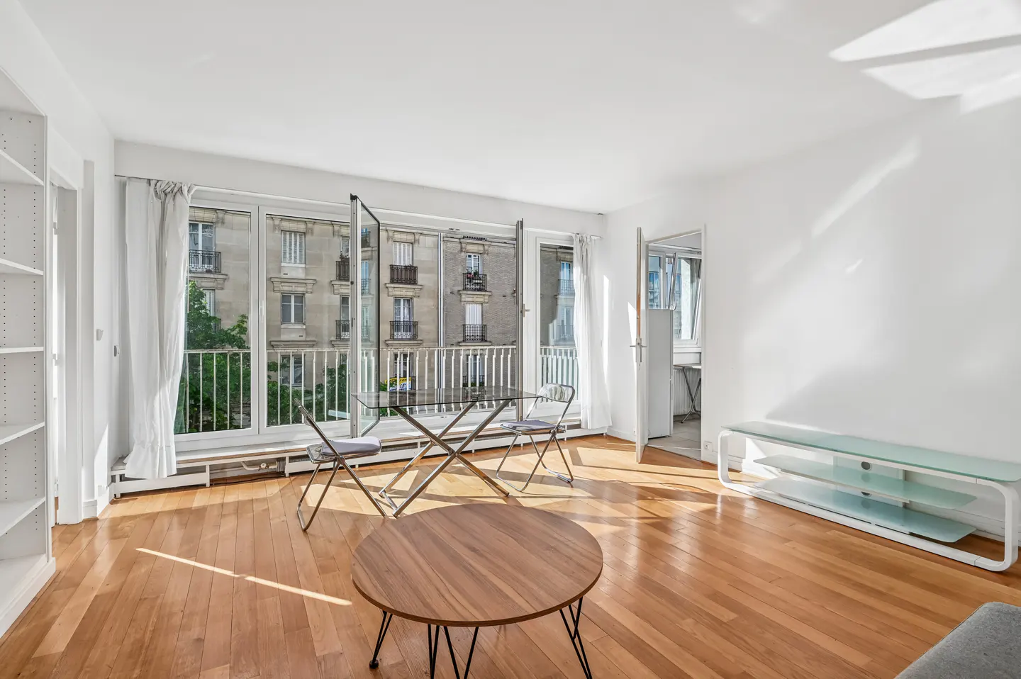 Bright, airy apartment interior with wood floors, white walls, and balcony view of a building across the street.