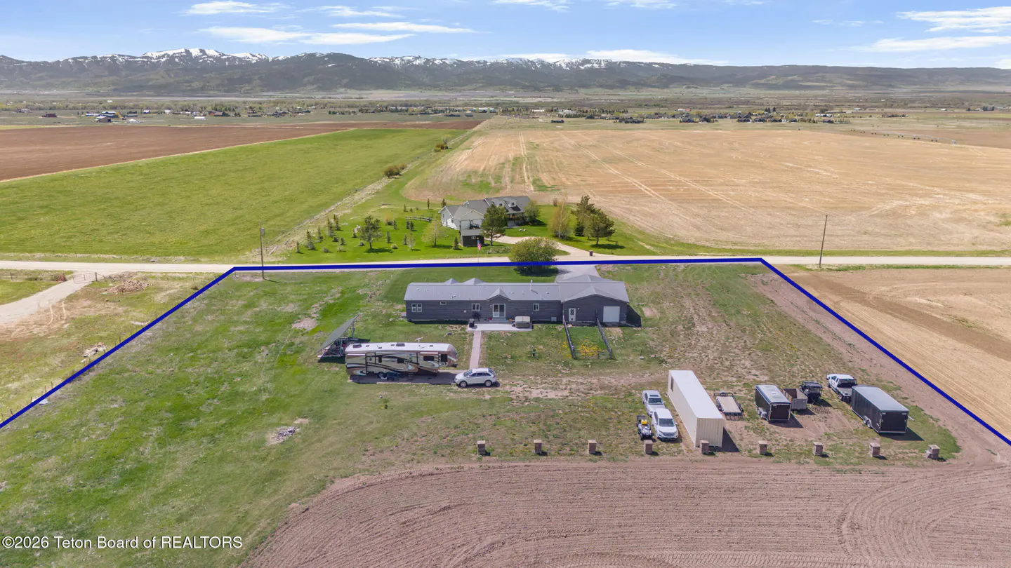 Aerial view of a gray ranch house with a blue property line, RV, vehicles, and trailers on a grassy lot with fields and mountains in the background.
